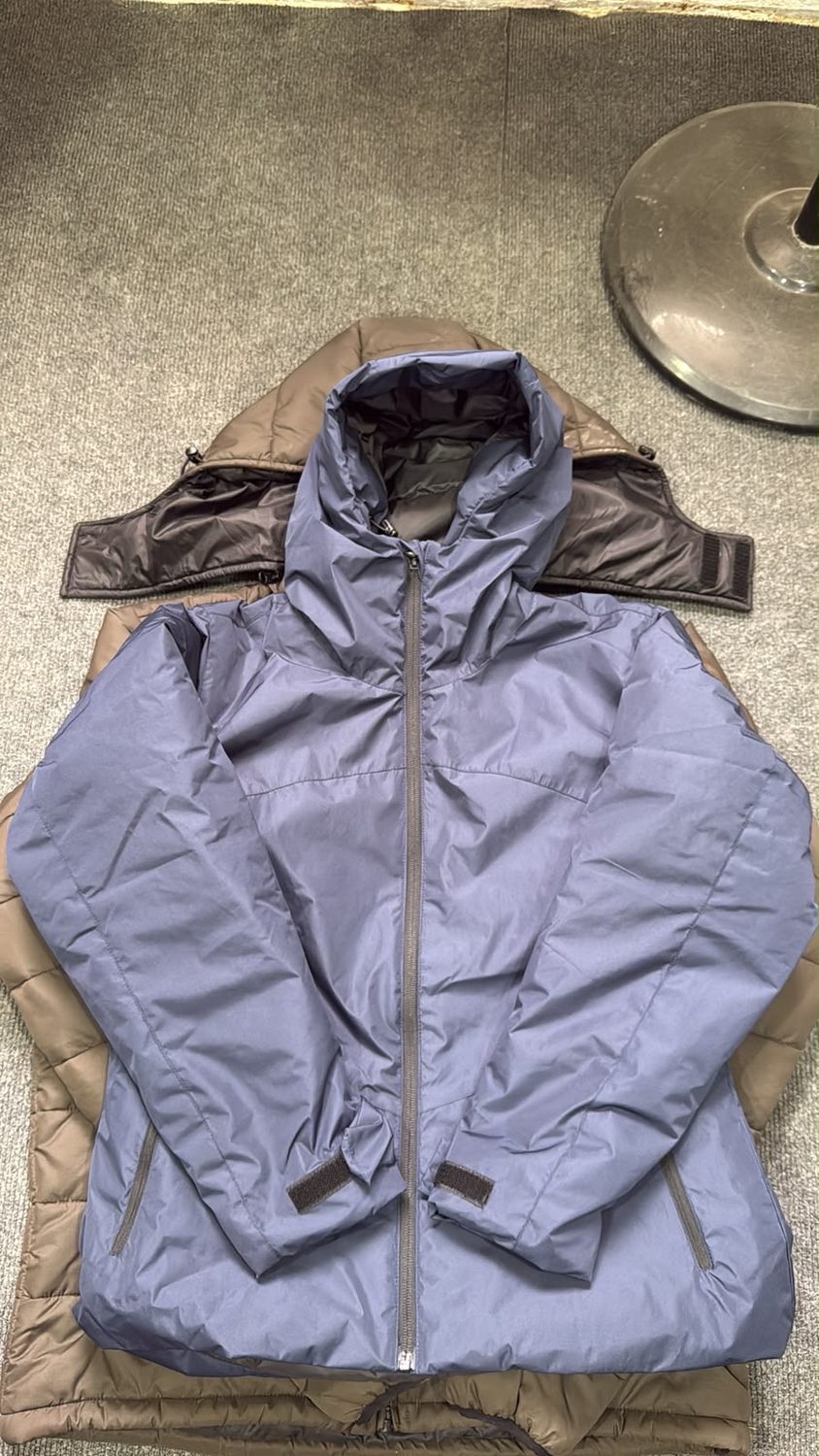 A periwinkle blue padded jacket laid over a tan jacket on grey carpet, with a round metal disc at the top right corner