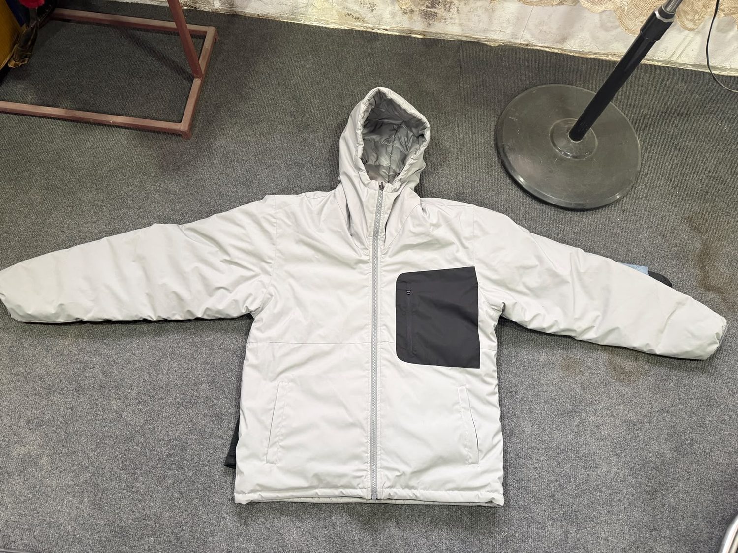 An off white hooded jacket laid flat on grey carpet with a large dark grey panel pocket on the chest