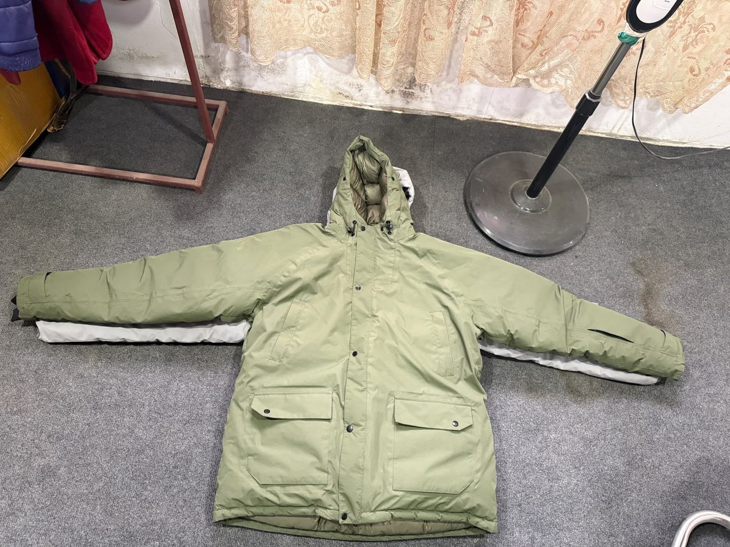 An olive hooded parka laid flat on grey carpet, showing a snap front placket, chest welt pockets and two flapped lower pockets