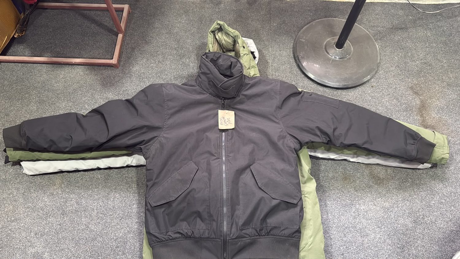 A charcoal jacket laid with both sleeves out on grey carpet over an olive jacket, a handwritten paper tag hanging from the front