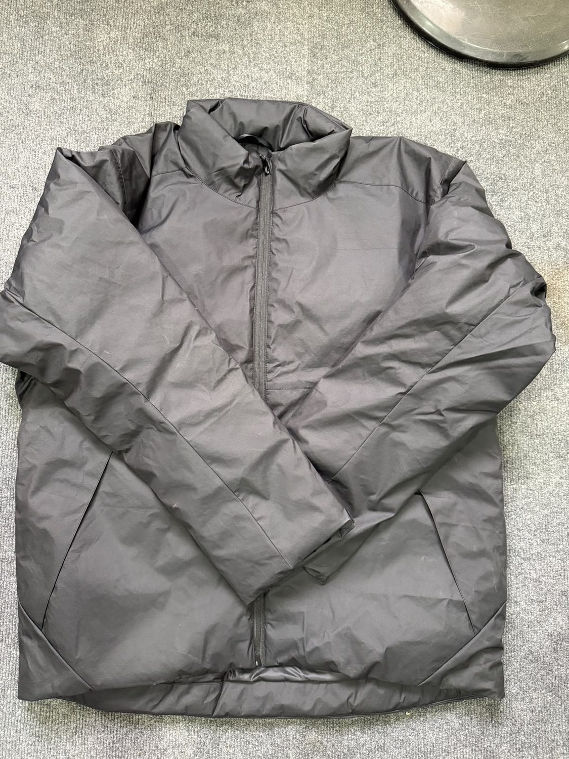 A dark grey jacket on grey carpet with a tall stand collar and no hood, front zip closed and sleeves folded across the body