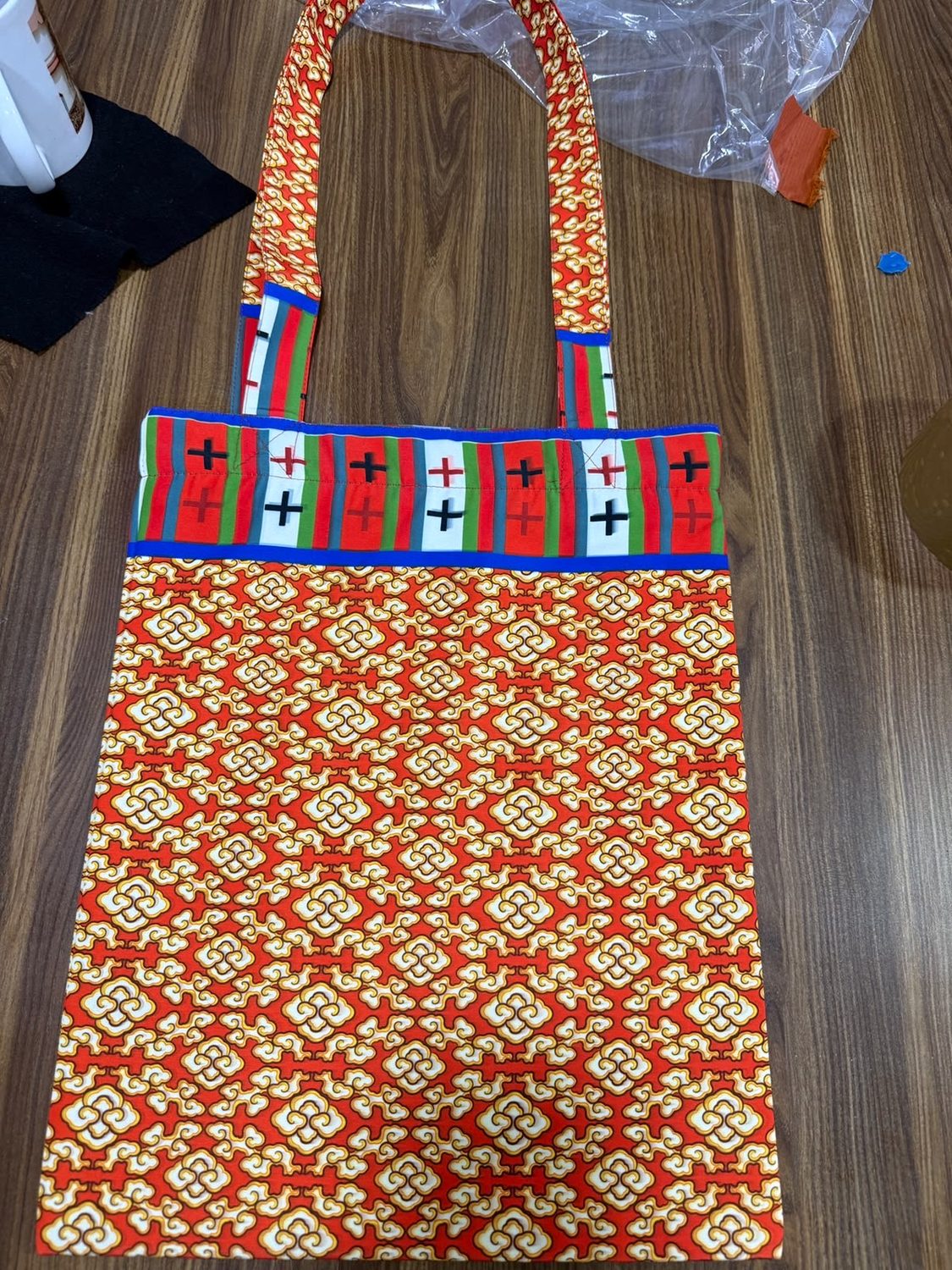 A flat tote bag of red cloth with a white and gold repeating motif and a striped woven band across the top