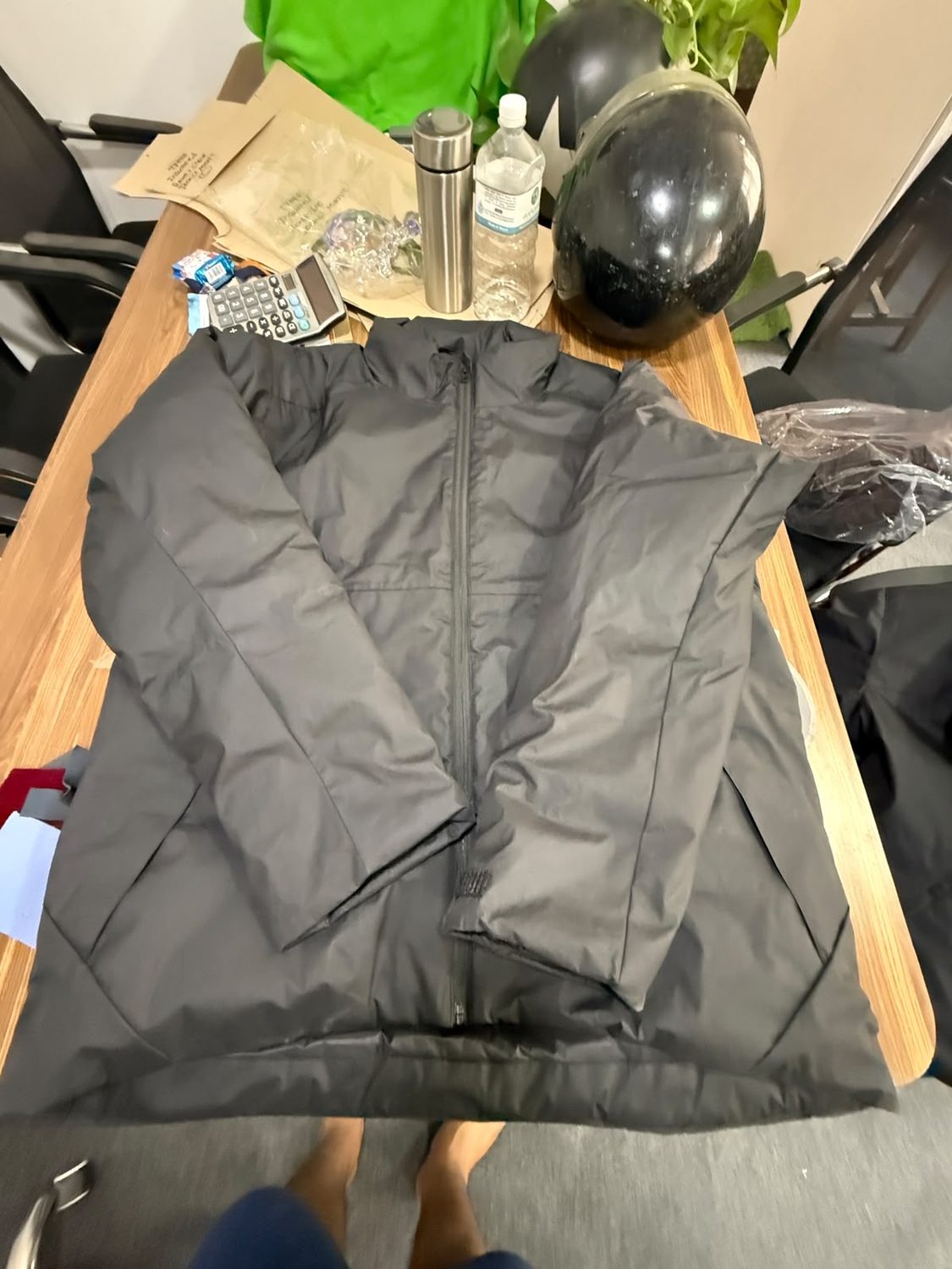A dark grey padded jacket lying open on a wooden workroom table with a calculator, a flask, a water bottle and a black helmet along the far edge